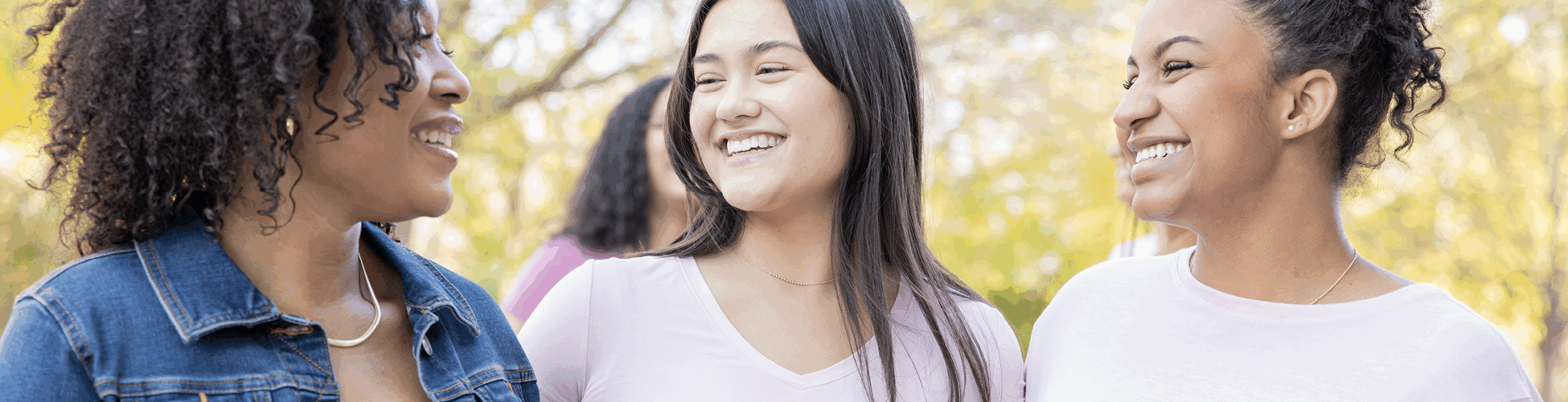 Three women wearing pink shirts with pink cancer bows attached smiling at each other.