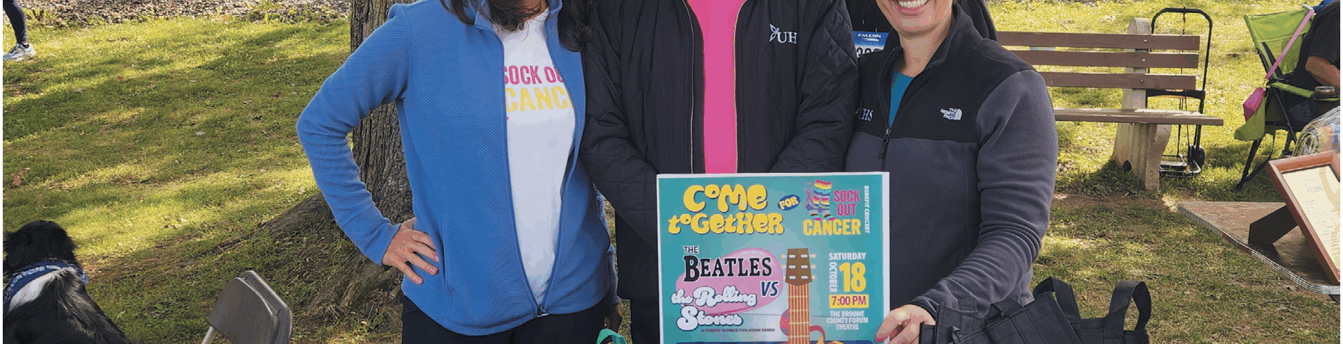 A man and two women stand together in the park in front of a table with goodie bags.