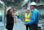 A woman and two men in hardhats talk, standing inside building under construction.