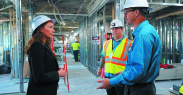 A woman and two men in hardhats talk, standing inside building under construction.