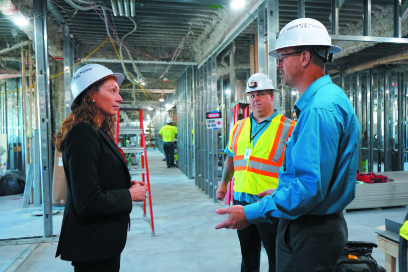 A woman and two men in hardhats talk, standing inside building under construction.
