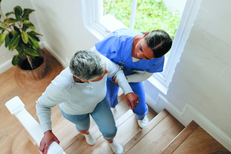 A home health worker helps an elderly woman walk up the stairs.